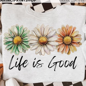 May include: A pale grey sweatshirt with three large daisy flowers in green, white, and orange with yellow centres. The words "Life is Good" are printed in black script below the flowers.