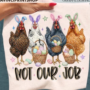 May include: A light pink t-shirt featuring four chickens wearing Easter bunny ears and holding baskets of Easter eggs. The text "NOT OUR JOB" is printed in a bold, black font with a yellow outline.