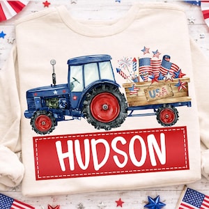 Könnte beinhalten: Cremefarbenes Sweatshirt mit einem blauen Traktor mit roten Rädern, der einen Holzkarren mit Feuerwerk und amerikanischen Flaggen zieht. Das Wort "USA" steht auf dem Wagen, und der Name "HUDSON" wird in einem roten Rechteck angezeigt.