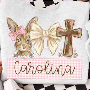 Puede incluir: Una sudadera gris claro con un diseño temático de Pascua. El diseño incluye un conejito con un lazo rosa, un lazo grande y una cruz. El nombre "Carolina" se muestra en una pancarta a cuadros rosa y blanco. Las palabras "DAVINCIPRINTSHOP" y "ORIGINAL DESIGNER" están impresas en la parte superior.