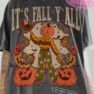 Pode incluir: Camiseta cinza escuro com o texto "IT'S FALL Y'ALL" e um gráfico de um cowboy com cabeça de abóbora. O gráfico inclui abóboras, morcegos e um desenho de corda. O cowboy usa um chapéu marrom, camisa amarela e calças verdes.