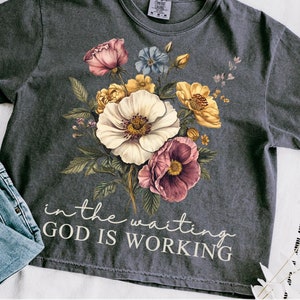May include: A dark gray t-shirt with a floral design and the text "in the waiting GOD IS WORKING".