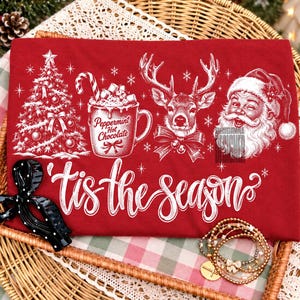 May include: A red t-shirt featuring a white graphic print of a Christmas tree, a mug labeled Peppermint Hot Chocolate, a reindeer with a bow, and Santa Claus. The text 'tis the season' is printed in white script below the holiday illustrations.