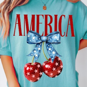 Könnte beinhalten: Ein türkisfarbenes T-Shirt mit dem Wort "AMERICA" in roten Buchstaben bedruckt. Ein blaues und weißes Band mit weißen Sternen befindet sich über zwei roten Kirschen mit weißen Sternen.