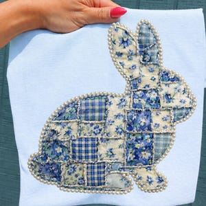 Puede incluir: Una toalla azul claro con un diseño de conejito de patchwork. El conejito está hecho de varias telas florales y a cuadros azules, delineadas con un ribete de perlas. La toalla está sostenida por una mano con esmalte de uñas rojo.