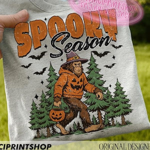 May include: Light gray sweatshirt with the words "SPOOKY Season" in orange and black. The graphic features a Bigfoot character wearing a pumpkin-themed hoodie and holding a jack-o'-lantern, with bats and pine trees. "DAVINCIPRINTSHOP" and "ORIGINAL DESIGNER" are printed at the bottom.