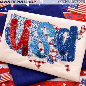 May include: A cream cushion with a map of the USA, the letters "USA" are in red and blue sequins. The map is made of various patriotic fabrics, including floral, gingham, and lace patterns. The cushion is set against a blue background with American flags.