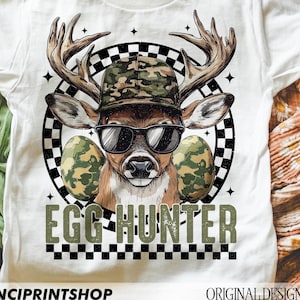 May include: White t-shirt featuring a deer wearing sunglasses and a camouflage hat, with the text "EGG HUNTER". The deer is surrounded by a black and white checkered pattern and two camouflage Easter eggs. A fun, festive design.