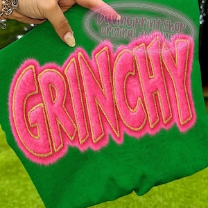 May include: Green fabric with the word "GRINCHY" in large, pink, fuzzy letters outlined in gold. The text is centered on the fabric. The words "DaVinciPrintShop original designer" are in a circle at the top.