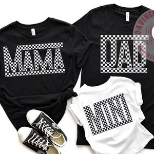 May include: Black t-shirts with white checkered text that reads "MAMA", "DAD", and "MINI". A pair of black sneakers and a white hat are also in the image. The shirts are likely family matching apparel.