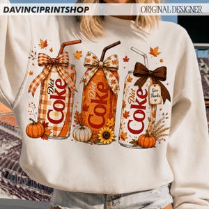 May include: A cream-colored sweatshirt featuring a graphic of three Diet Coke cans decorated with autumn themes. Each can has a straw, a bow, and fall elements like pumpkins, sunflowers, and leaves. One can has a tag that says Give Thanks.