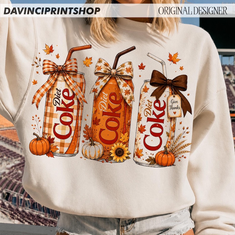 May include: A cream-colored sweatshirt featuring a graphic of three Diet Coke cans decorated with autumn themes. Each can has a straw, a bow, and fall elements like pumpkins, sunflowers, and leaves. One can has a tag that says Give Thanks.