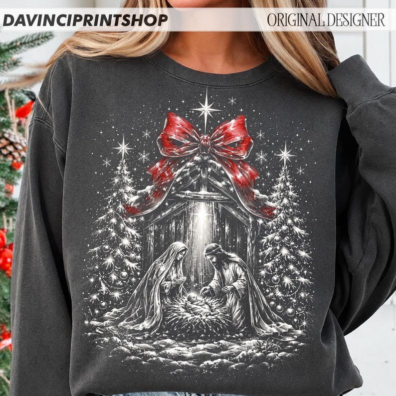 May include: A charcoal gray crewneck sweatshirt featuring a white graphic print of a nativity scene with two figures kneeling over a manger. A large red bow sits above the scene, flanked by two pine trees under a bright star and snowflakes.