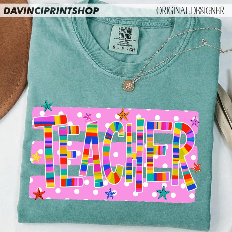 May include: A sage green crew neck t-shirt featuring a graphic print of the word TEACHER in multicolored stripes. The design includes a pink rectangular background with white polka dots and small colorful star accents. A silver necklace rests on top.