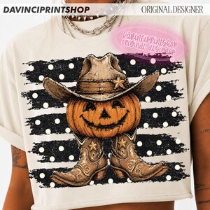 K&ouml;nnte beinhalten: Ein cremefarbenes T-Shirt mit einer Grafik eines orangefarbenen K&uuml;rbisses, der einen braunen Cowboyhut und braune Cowboystiefel tr&auml;gt. Das Design ist vor einem Hintergrund aus schwarzen horizontalen Streifen mit wei&szlig;en Punkten platziert.