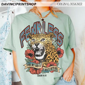 Puede incluir: Camiseta verde claro con un dise&ntilde;o gr&aacute;fico. El dise&ntilde;o incluye la palabra "FEARLESS" en rojo y azul, un leopardo y flores rojas. Tambi&eacute;n se incluye el texto "FOR MY GOD IS ALWAYS WITH ME" e "ISAIAH 41:10".