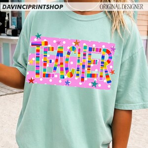 May include: A light mint green short-sleeve t-shirt featuring a rectangular pink graphic with white polka dots. The word TEACHER is printed in large, multi-colored striped letters, surrounded by small, colorful, patterned star shapes.