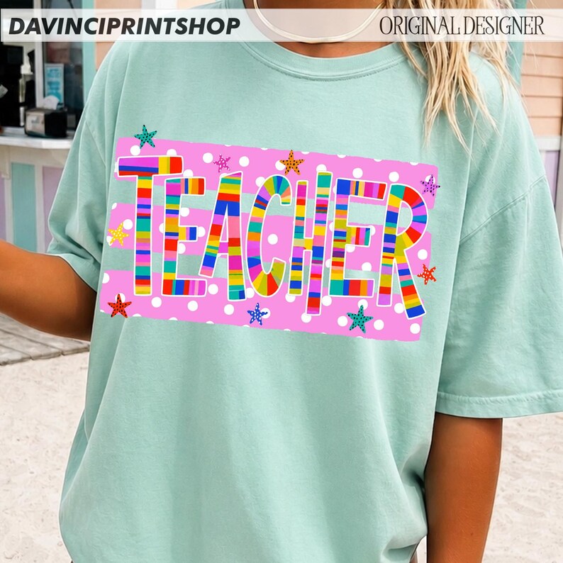 May include: A light mint green short-sleeve t-shirt featuring a rectangular pink graphic with white polka dots. The word TEACHER is printed in large, multi-colored striped letters, surrounded by small, colorful, patterned star shapes.