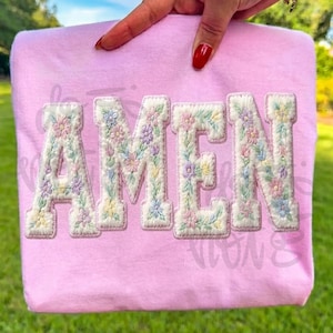 Könnte beinhalten: Ein hellrosa Sweatshirt mit dem Wort "AMEN" in großen, floral verzierten Buchstaben. Die Buchstaben sind weiß mit pastellfarbenen Blumen. Das Sweatshirt wird von einer Hand mit rotem Nagellack gehalten, vor einem Hintergrund aus grünem Gras.