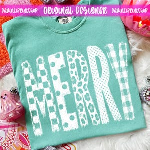 May include: A teal crewneck t-shirt featuring the word MERRY printed in large white letters. Each letter displays a different pattern, including checkers, polka dots, and floral motifs. The shirt has a Comfort Colors brand label at the neck.