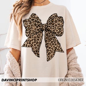 May include: A beige t-shirt with a large black and brown leopard print bow design. The shirt has short sleeves and a crew neck.