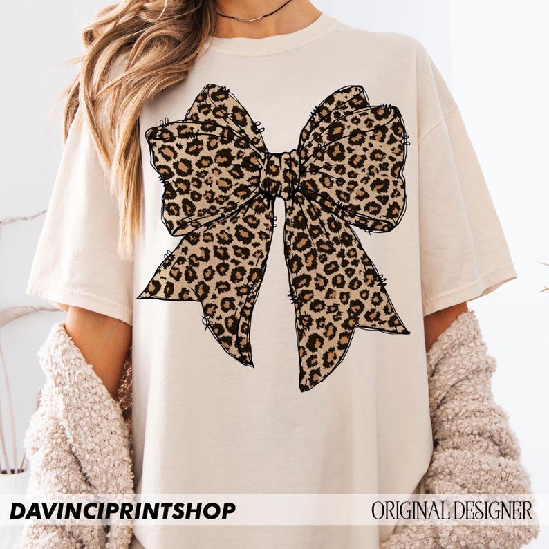 May include: A beige t-shirt with a large black and brown leopard print bow design. The shirt has short sleeves and a crew neck.