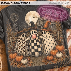 K&ouml;nnte beinhalten: Ein anthrazitfarbenes T-Shirt mit einer Grafik von drei Geistern mit Hexenh&uuml;ten auf einem Feld mit K&uuml;rbissen, einer Windm&uuml;hle und einem Haus unter einem Vollmond. Die Geister haben Blumen-, Karomuster und Punkte. Der Text lautet DAVINCIPRINTSHOP.