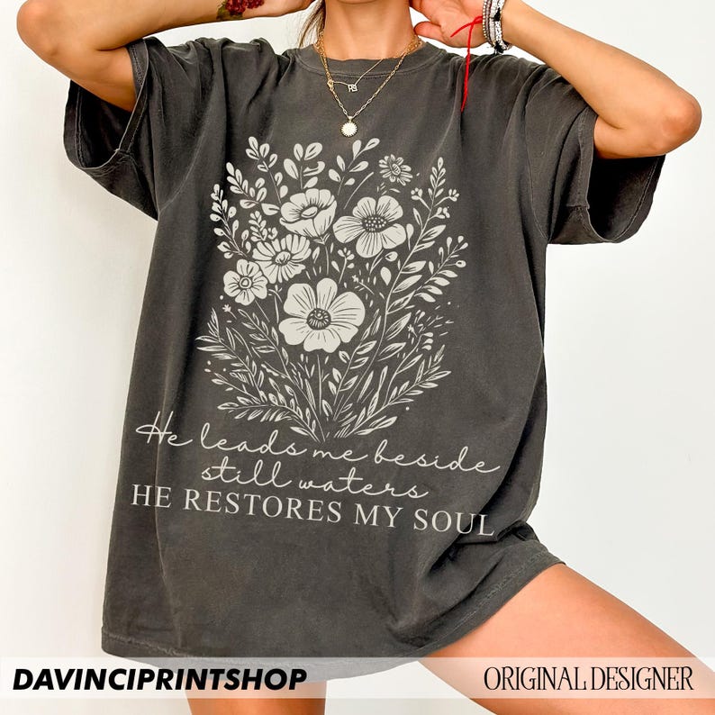 May include: A dark gray oversized t-shirt with a white floral design and the text "He leads me beside still waters HE RESTORES MY SOUL".