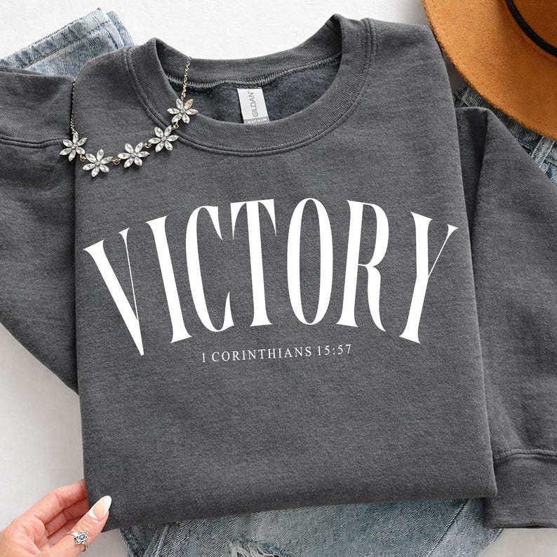 K&ouml;nnte beinhalten: Dunkelgraues Sweatshirt mit dem Wort "VICTORY" in gro&szlig;en wei&szlig;en Buchstaben. Darunter steht der Text "1 Korinther 15,57". Eine silberne Blumenkette liegt auf dem Sweatshirt.
