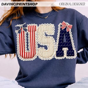 May include: Navy blue sweatshirt with a patriotic "USA" design. The letters are embellished with red and white stripes, a cable knit texture, and a navy blue star pattern. Each letter is outlined with a white lace trim, and topped with a bow.