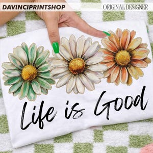 May include: White t-shirt with three large daisy flowers in green, white, and orange with yellow centres. The phrase "Life is Good" is written in black script below the flowers. The shirt is displayed on a green and white checkered surface.