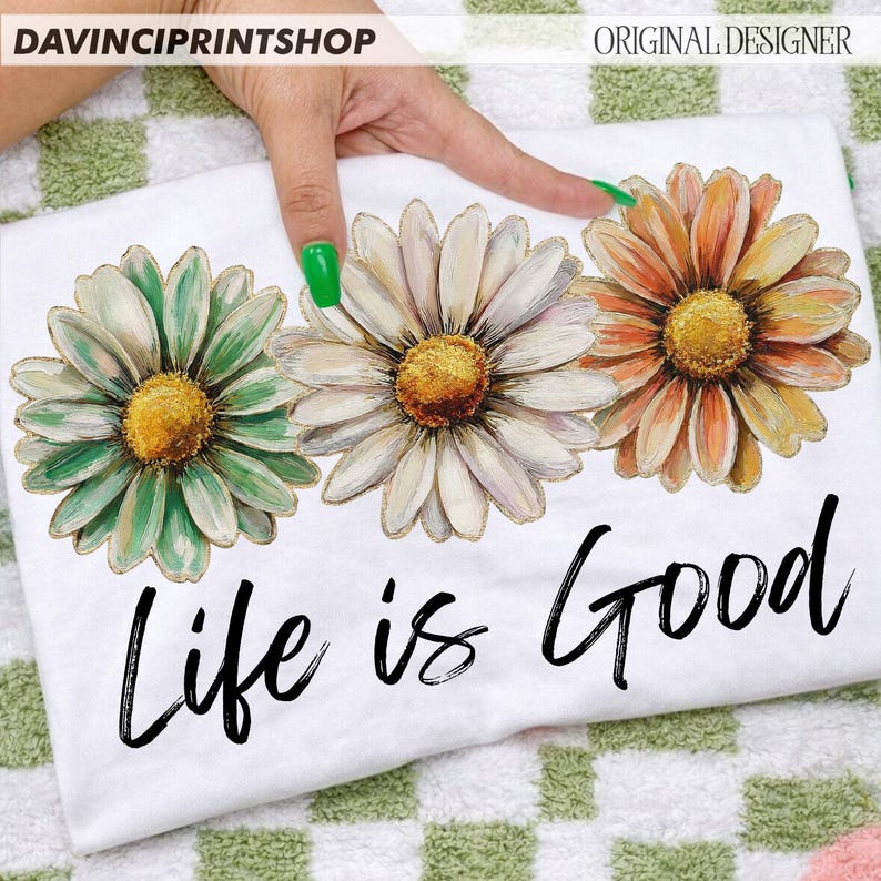 May include: White t-shirt with three large daisy flowers in green, white, and orange with yellow centres. The phrase "Life is Good" is written in black script below the flowers. The shirt is displayed on a green and white checkered surface.