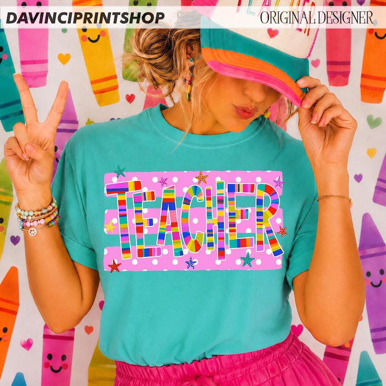 May include: A teal t-shirt featuring the word TEACHER in colorful, multi-striped block letters centered on a pink rectangular background with white polka dots and small star accents. The shirt is worn with a pink and teal baseball cap.