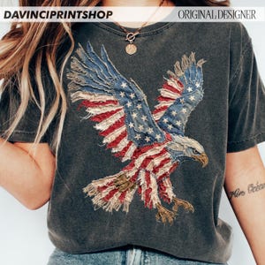 Pu&ograve; includere: T-shirt grigio scuro con un design a forma di aquila della bandiera americana invecchiata. Le ali dell'aquila sono realizzate in tessuto blu con stelle bianche e strisce rosse e bianche. La maglietta ha il testo "DAVINCIPRINTSHOP" e "ORIGINAL DESIGNER".