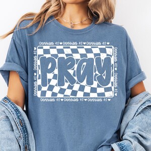 Op de afbeelding: Een blauw T-shirt met een witte geruite achtergrond en het woord "PRAY" in witte letters. De tekst "Kolossenzen 4:2" wordt herhaald rond de geruite achtergrond.