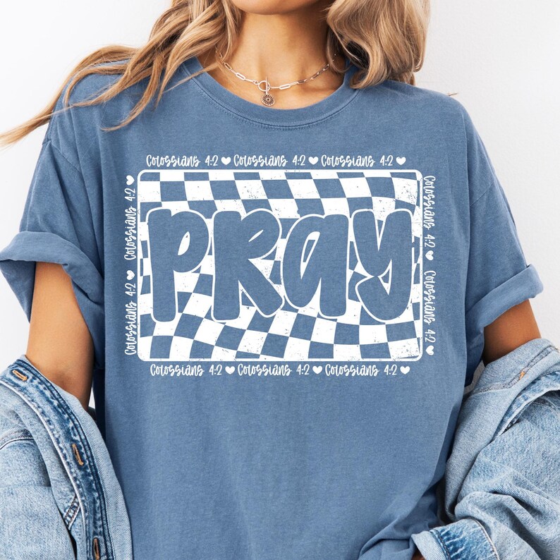 Op de afbeelding: Een blauw T-shirt met een witte geruite achtergrond en het woord "PRAY" in witte letters. De tekst "Kolossenzen 4:2" wordt herhaald rond de geruite achtergrond.