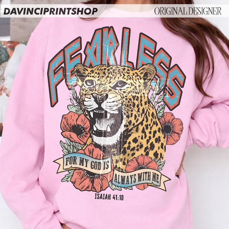 Puede incluir: Sudadera rosa claro con un leopardo rugiente, la palabra "FEARLESS" en verde azulado y naranja, y el texto "FOR MY GOD IS ALWAYS WITH ME" con el vers&iacute;culo b&iacute;blico Isa&iacute;as 41:10. El dise&ntilde;o incluye flores rojas.