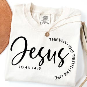 May include: A beige t-shirt with black text that reads "Jesus" and "John 14:6". The text is in a circle with the words "The Way, The Truth, The Life" around the outside.