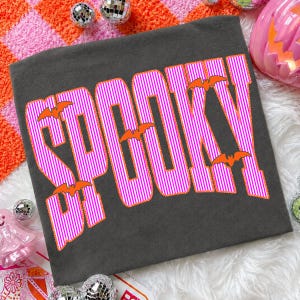 May include: A dark gray t-shirt featuring the word SPOOKY printed in large, bold letters. The letters have a vertical pink and white striped pattern with a bright orange outline. Several small, solid orange bat silhouettes are placed around the text.