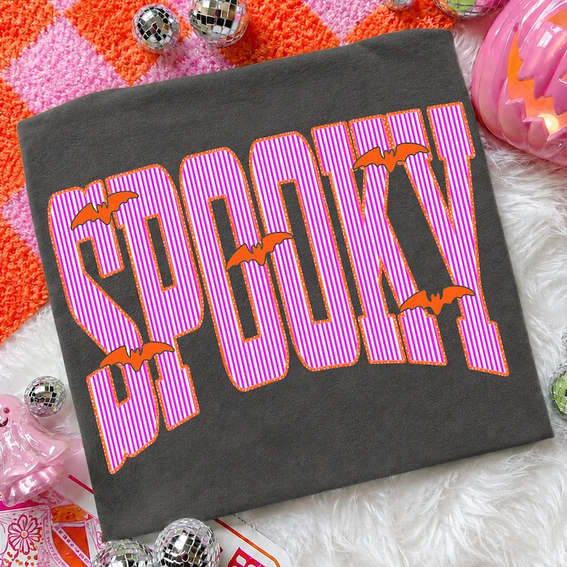 May include: A dark gray t-shirt featuring the word SPOOKY printed in large, bold letters. The letters have a vertical pink and white striped pattern with a bright orange outline. Several small, solid orange bat silhouettes are placed around the text.