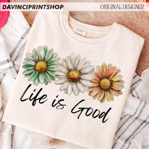 May include: A cream-coloured sweatshirt displaying three daisy flowers in green, white, and orange with yellow centres. The words "Life is Good" are printed in black script below the flowers. The top is from Davinci Printshop.