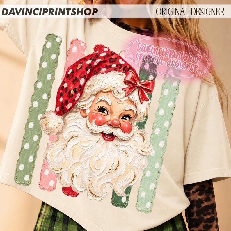 May include: A cream-colored t-shirt featuring a graphic of a Santa Claus face with a red leopard print hat and a red bow. The design includes two vertical green stripes with white polka dots on either side of the Santa illustration.