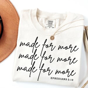 Pode incluir: Uma camiseta bege com o texto "made for more" repetido tr&ecirc;s vezes em escrita cursiva preta. O texto "Ephesians 2:10" &eacute; impresso em preto abaixo da escrita cursiva.