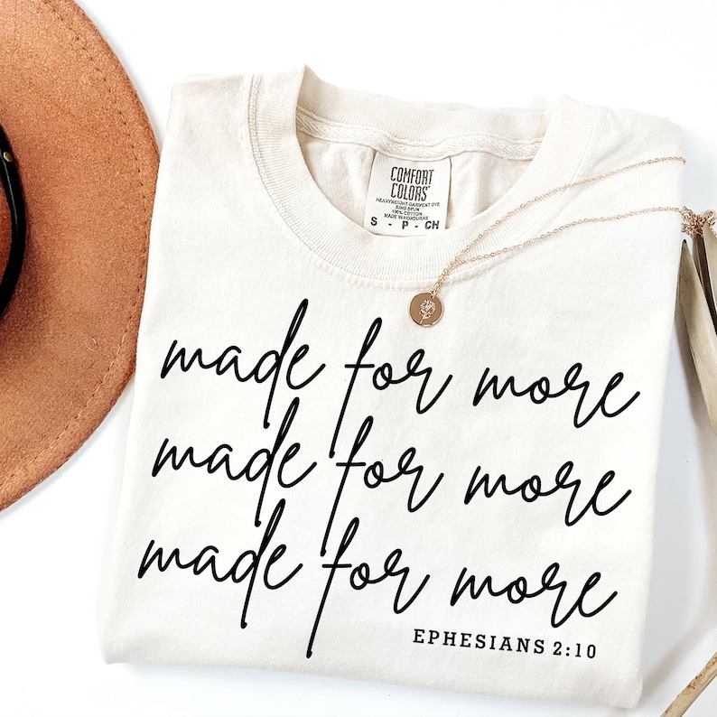 Pode incluir: Uma camiseta bege com o texto "made for more" repetido tr&ecirc;s vezes em escrita cursiva preta. O texto "Ephesians 2:10" &eacute; impresso em preto abaixo da escrita cursiva.