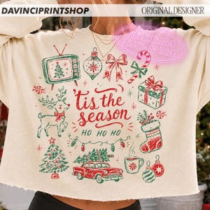 May include: A cream-colored crewneck sweatshirt featuring a festive red and green graphic print. The design includes a vintage television, reindeer, Christmas tree, gift box, stocking, candy cane, and a car, with the text 'tis the season HO HO HO'.