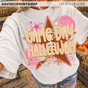May include: A white short-sleeve t-shirt featuring a graphic print of a brown star, a pink football helmet, and pink paint splatters. The text "GAME DAY HALLELUJAH" is printed in large, light tan letters with a white outline across the center.