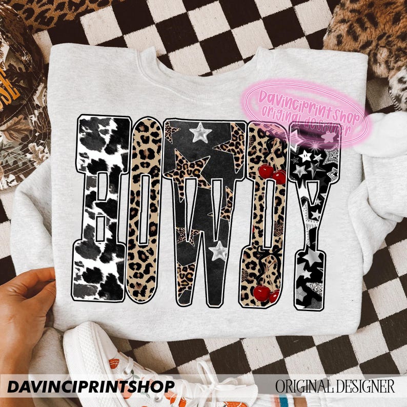May include: A light gray sweatshirt with the word "HOWDY" in a bold, decorative font. The letters feature a mix of patterns, including cow print, leopard print, and stars. The text "DAVINCIPRINTSHOP" is printed at the bottom.