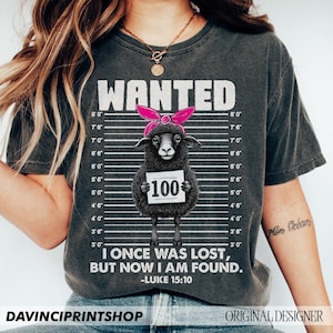 Puede incluir: Una camiseta negra con un aspecto desgastado presenta una oveja negra que lleva un pa&ntilde;uelo rosa y sostiene un cartel que dice "100". La oveja est&aacute; rodeada de un fondo a rayas blanco y negro con medidas de altura. El texto "WANTED" est&aacute; en la parte superior de la camiseta. Debajo de la oveja, el texto dice "I once was lost, but now I am found. -Luke 15:10".
