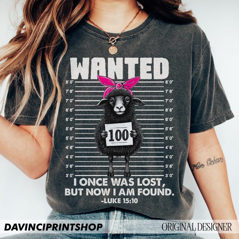 Puede incluir: Una camiseta negra con un aspecto desgastado presenta una oveja negra que lleva un pa&ntilde;uelo rosa y sostiene un cartel que dice "100". La oveja est&aacute; rodeada de un fondo a rayas blanco y negro con medidas de altura. El texto "WANTED" est&aacute; en la parte superior de la camiseta. Debajo de la oveja, el texto dice "I once was lost, but now I am found. -Luke 15:10".