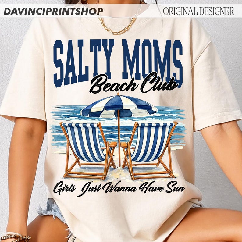 Op de afbeelding: Cr&egrave;mekleurig T-shirt met de tekst "Salty Moms Beach Club" in blauw en zwart. Het ontwerp toont een strandsc&egrave;ne met blauw-wit gestreepte stoelen, een blauw-witte parasol en de zin "Girls Just Wanna Have Sun."